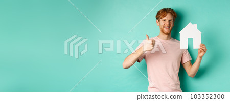 Real estate concept. Young man with red hair, wearing t-shirt, showing paper house cutout and thumb up, recommend agency, standing over turquoise background 103352300