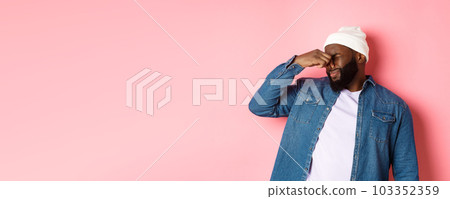 Disgusted african-american man shut nose from aversion, displeased by awful bad smell, standing in beanie and denim shirt over pink background 103352359