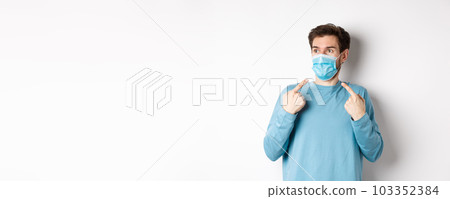 Coronavirus, health and quarantine concept. Confused young man pointing at medical mask on face and looking left, standing over white background 103352384