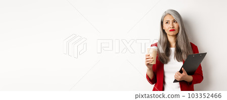 Sad asian businesswoman drinking coffee at work and looking upper left corner, standing over white background Sad asian businesswoman drinking coffee at work and looking upper left corner, standing over white background 103352466