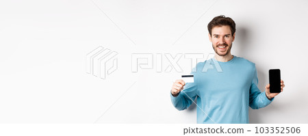 Smiling caucasian man showing plastic credit card with mobile phone screen. Guy recommending online banking app, standing on white background 103352506