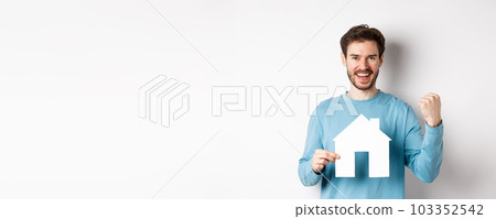 Real estate and insurance concept. Cheerful man buying property and celebrating, saying yes and showing paper house cutout, standing on white background 103352542