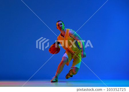 Male basketball player in yellow uniform during game, dribbling ball against blue studio background in neon light. Male basketball player in yellow uniform during game, dribbling ball against blue studio background in neon light. 103352725
