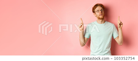 Dreamy young man with red hair and beard pointing fingers up, looking pensive at top, standing against pink background 103352754