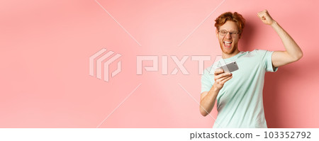 Happy redhead man winning in mobile video game, raising hand up and shouting yes with joy, celebrating victory, looking at smartphone, standing over pink background Happy redhead man winning in mobile video game, raising hand up and shouting yes with joy, celebrating victory, looking at smartphone, standing over pink background 103352792