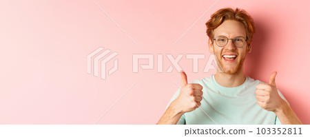 Face of satisfied male customer showing thumbs-up in approval, smiling happy, wearing glasses and t-shirt, pink background 103352811