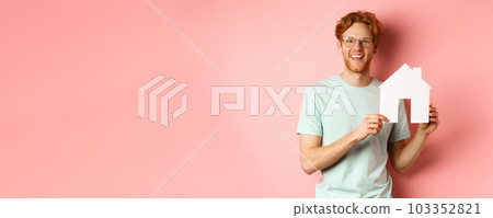 Real estate. Handsome redhead man in t-shirt and glasses, showing paper house cutout and smiling, standing over pink background Real estate. Handsome redhead man in t-shirt and glasses, showing paper house cutout and smiling, standing over pink background 103352821