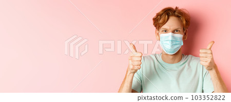 Covid-19 and pandemic concept. Handsome guy with messy ginger hair, wearing medical mask on face and showing thumbs up, standing over pink background Covid-19 and pandemic concept. Handsome guy with messy ginger hair, wearing medical mask on face and showing thumbs up, standing over pink background 103352822