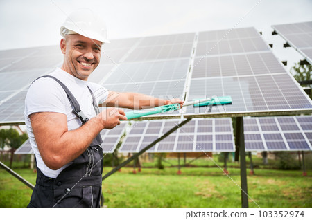 Professional worker cleaning solar panel from dust. Smiling man wiping PV battery using mop. Man holding mop and smiling straight to camera. Professional worker cleaning solar panel from dust. Smiling man wiping PV battery using mop. Man holding mop and smiling straight to camera. 103352974