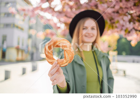 Attractive young female tourist photographer with stylish clothes and a boho hat holds bagel obwarzanek traditional polish cuisine snack walking around on blossoming sakura trees background in Krakow 103352990