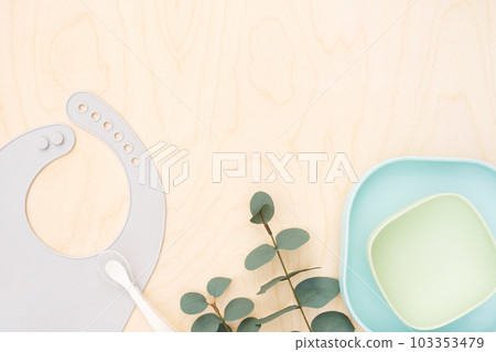 Baby accessories and tableware for food on wooden background. First feeding for kids concept. Flat lay, Top View, Space for text. Silicone dishware, bib, cup on neutral backdrop. Serving baby food 103353479