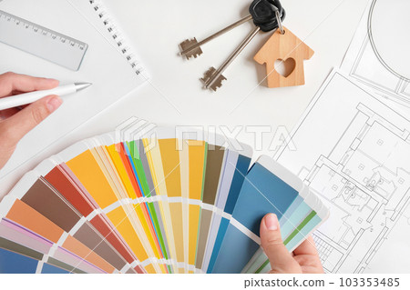 Hands choosing Colors of Interior Design from the Color Guide. Apartment Sketch, Paint palette, Model of House and Keyring on the Background. Architect Using Color Swatch. House Plan with Blueprints 103353485