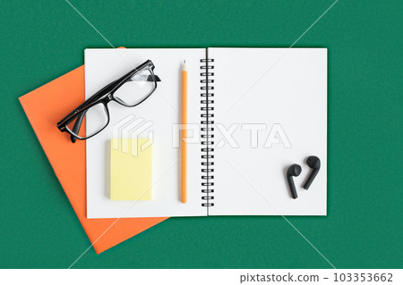 Notebook with pencil, glasses, wireless headphones. Open notebook on dark green color background, spiral notepad. Office desk. Copy Space Notebook with pencil, glasses, wireless headphones. Open notebook on dark green color background, spiral notepad. Office desk. Copy Space 103353662