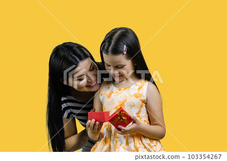 Little girl is opening a red gift box together with her mom. Isolated on yellow background. 103354267