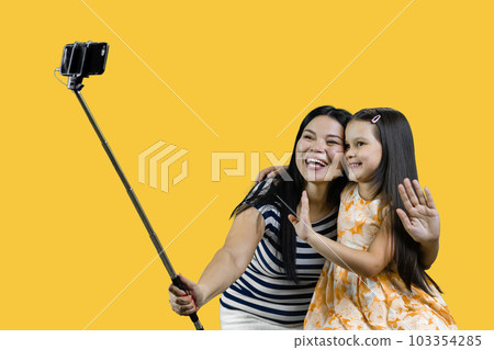 Mom and her daughter are making a common photo on smartphone using selfie stick. Isolated on yellow background. 103354285