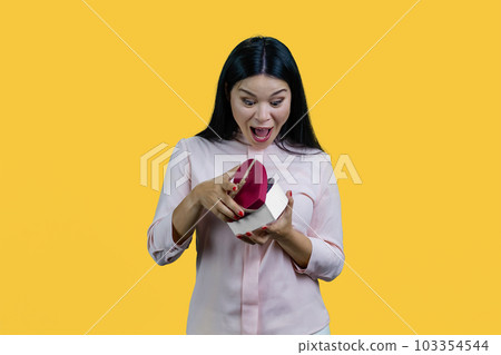 Extremely happy and surprised asian woman opening heart shape gift box. Valentines day or anniversary. Isolated on yellow background. Extremely happy and surprised asian woman opening heart shape gift box. Valentines day or anniversary. Isolated on yellow background. 103354544