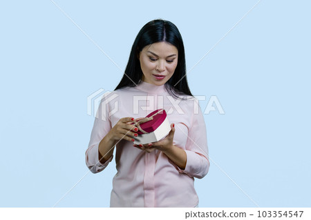 Portrait of a young asian woman opens a heart shape gift box. Isolated on pale blue background. Portrait of a young asian woman opens a heart shape gift box. Isolated on pale blue background. 103354547