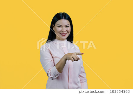 Portrait of happy cheerful asian woman is pointing at copy space. Isolated on yellow background. 103354563