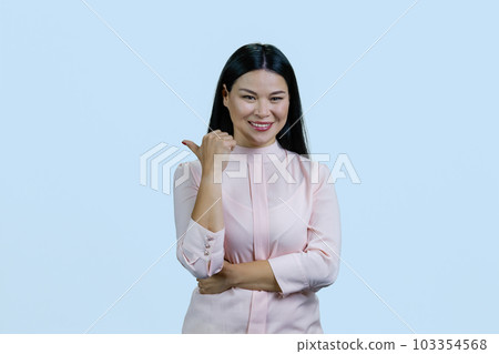 Portrait of happy cheerful asian woman is pointing back with her thumb. Isolated on pastel blue background. 103354568