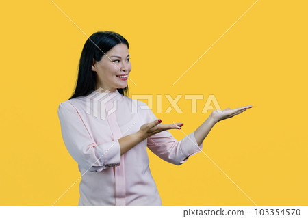 Happy young asian woman presenting copy space with both hands. Isolated on yellow background. 103354570