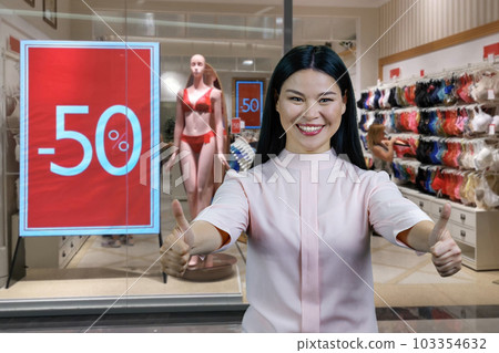 Happy young asian woman shows both thumb up. Lingerie boutique with half discount in the background. Happy young asian woman shows both thumb up. Lingerie boutique with half discount in the background. 103354632