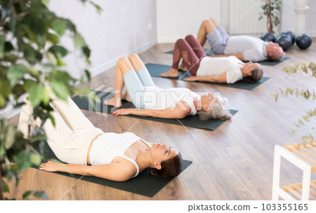 Class of elderly sporty women stretching gluteal muscle while lying on back during yoga Pilates workout on mats in gym studio Class of elderly sporty women stretching gluteal muscle while lying on back during yoga Pilates workout on mats in gym studio 103355165