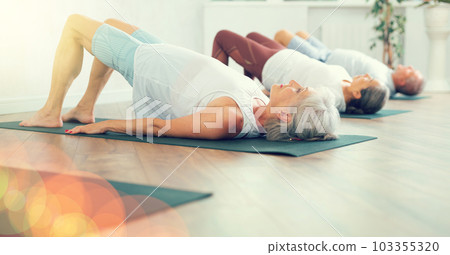 Elderly woman in company of hatha yoga lovers performs bridge pose, setu bandha sarvangasana 103355320