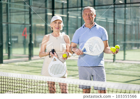 Old man and woman posing with Padel racket in hands next to the net in tennis court 103355566