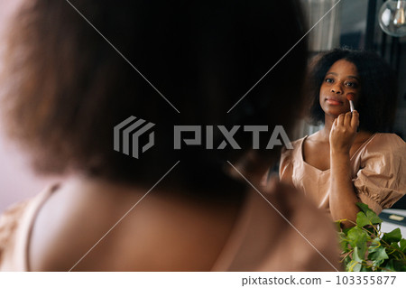 Closeup rear view from shoulder of attractive African young woman using cosmetics brush, looking to reflection on mirror. Beautiful black female putting daily makeup, applying powder at home. 103355877