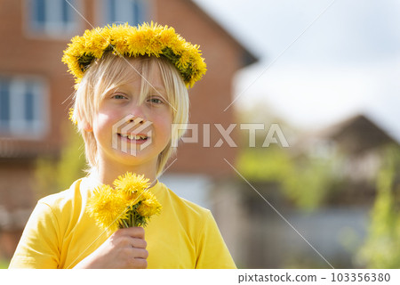 Portrait of Caucasian blond boy with wreath of dandelions and bouquet of flowers near the house. Blurred background. 103356380