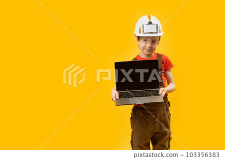 Schoolboy in work clothes and protective helmet holds laptop with empy screen. Child as architect. Choice of future profession. Copy space. Schoolboy in work clothes and protective helmet holds laptop with empy screen. Child as architect. Choice of future profession. Copy space. 103356383
