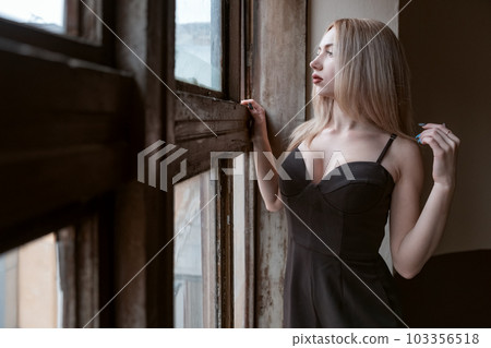 Portrait of attractive young woman in black revealing outfit with deep neckline stands near an old window and looks out outside. 103356518