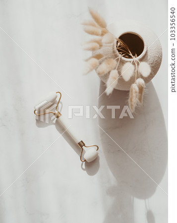 White Jade face roller for beauty facial massage therapy and dry flowers in vase on white background with shadows 103356540
