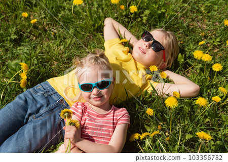 Portrait of two blonde children in sunglasses lying on dandelions field. Little brother and sister basking in the sun in meadow. Portrait of two blonde children in sunglasses lying on dandelions field. Little brother and sister basking in the sun in meadow. 103356782