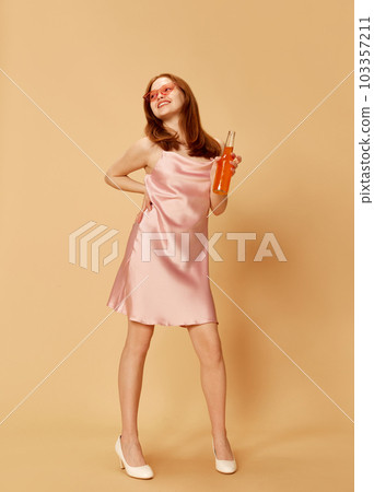 Beautiful woman with red hair wearing elegant pink dress holding drink bottle and posing with happy smiling face over orange studio background. Concept of party, youth 103357211