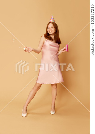 Cheerful young girl, redhead woman wearing dress and birthday paper cone using bubble blower and smiling over orange studio background 103357219