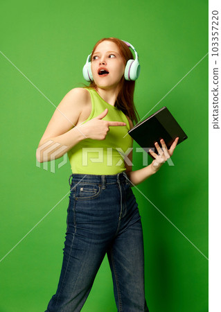 One young girl, student wearing headphones holding tablet and pointing on it over green studio background. Concept of emotions, happiness One young girl, student wearing headphones holding tablet and pointing on it over green studio background. Concept of emotions, happiness 103357220
