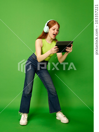 Emotive young girl, a teenager wearing headphones holding a tablet and looking at the screen, playing with interested face. Video games 103357221