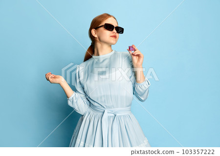 Portrait with young lady, woman with foxy hair wearing dress and black sunglasses posing with smile over blue background. Concept of medieval era, beauty, modern fashion Portrait with young lady, woman with foxy hair wearing dress and black sunglasses posing with smile over blue background. Concept of medieval era, beauty, modern fashion 103357222