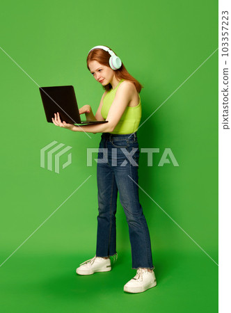 One young girl, student wearing headphones holding laptop and looking at screen with serious face. Concept of emotions, happiness, student life. Online education 103357223