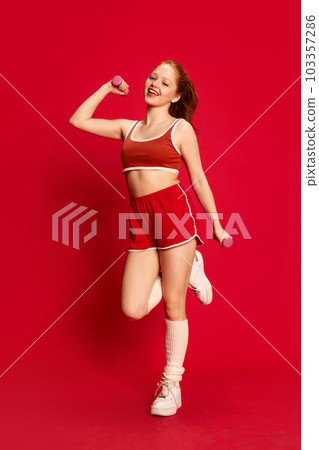Cheerful young girl wearing sportswear doing sport exercises with pleasure over red background. Female fitness 103357286