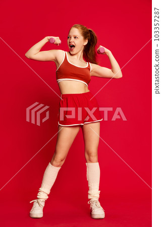 Charming red haired young girl wearing sportswear training with dumbbells with happy face on red background. Sport workout for slim body 103357287