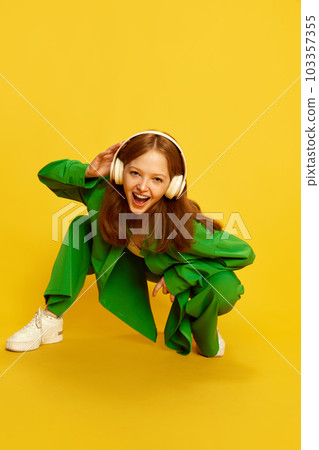 Funny girl, teenager with red hair wearing wireless headphones listening to music over yellow studio background. Top view. Concept of youth, fashion 103357355