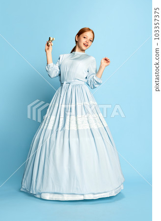 Portrait with one charming girl, princess with red hair wearing festive dress holding theatre binoculars smiling and posing over blue background. Concept of medieval era, beauty 103357375