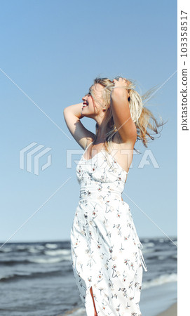 Happy blonde woman in free happiness bliss on ocean beach standing straight. Portrait of a female model in white summer dress enjoying nature during travel holidays vacation outdoors 103358517