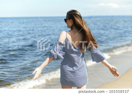 Happy smiling woman in free bliss on ocean beach standing with open hands. Portrait of a brunette female model in summer dress enjoying nature during travel holidays vacation outdoors, back view 103358537