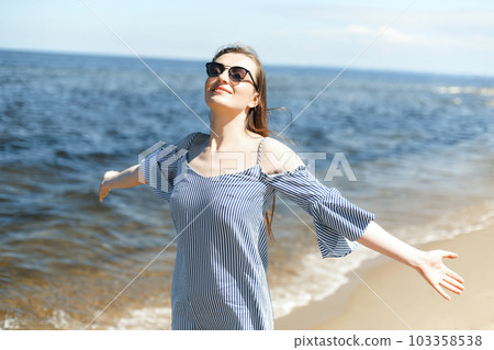 Happy smiling woman in free bliss on ocean beach standing with open hands. Portrait of a brunette female model in summer dress enjoying nature during travel holidays vacation outdoors Happy smiling woman in free bliss on ocean beach standing with open hands. Portrait of a brunette female model in summer dress enjoying nature during travel holidays vacation outdoors 103358538