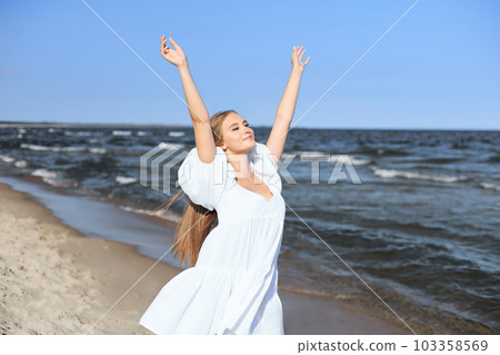 Happy smiling beautiful woman on the ocean beach standing in a white summer dress, raising hands Happy smiling beautiful woman on the ocean beach standing in a white summer dress, raising hands 103358569