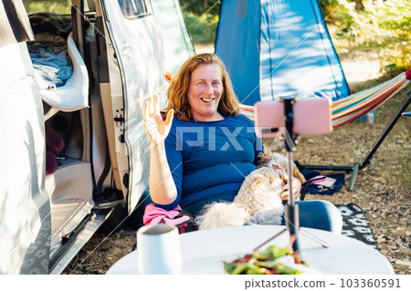 Middle age nomad woman having video call on phone while sitting with dog pet near camper van in forest camp. Travel blogger working during road trip vacation. Online communication with followers. Middle age nomad woman having video call on phone while sitting with dog pet near camper van in forest camp. Travel blogger working during road trip vacation. Online communication with followers. 103360591