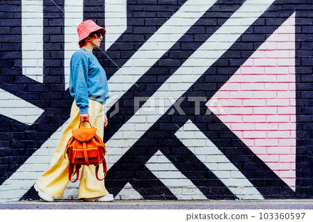 Hipster fashion young woman in bright clothes, sun glasses, bucket hat with backpack walking on the background of the painted brick wall. Urban city street fashion. Selective focus. Copy space. 103360597